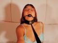 AlyssaGreen with glasses, brown hair, wearing a black ball gag and blue lace bra, eyes closed, black leash attached to collar, tan skin, sitting against white wall.