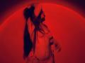 Red-lit photo of Tisia with long hair, wearing a pink bra and panties, her tits and ass slightly visible, arms raised, hands near her head, in a sensual pose.