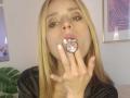 LyannaDifran, blonde, with pink-tinted lips, sucking a pink butt plug, hand with white nail polish, wearing black top, bedroom setting, light skin, brown eyes, subtle makeup.
