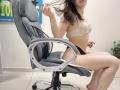 JoannaSia, de piel pálida, cabello castaño, con senos pequeños en un sujetador de encaje, ropa interior negra, sentada en una silla de oficina gris, sujetándose el cabello, suelo de baldosas blancas, reposabrazos grises, planta en maceta..