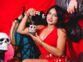 LilyConorr with black hair, red dress, and gold bracelets holds a skull jar, sitting on a gold chair, next to a skull and black feathers, smiling. Her tits are visible.