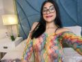 NinaRouse with long black hair, glasses, and red lipstick, wearing a sheer, colorful, geometric-patterned top, exposing her tits and nipples, smiling in a brightly lit bedroom.