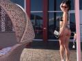 Milagres with tan skin, dark hair, and sunglasses, wearing a black bra and leopard print thong, standing with her ass facing the camera, holding a black bag. Wicker chair.
