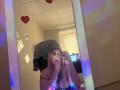DiLaria with blonde hair, holding phone with rainbow lights, covering face, showing large tits, tattoo on chest, mirror selfie, wearing black strap, gray shirt on wall.