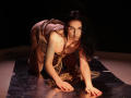 TriciaBarnes on all fours, wearing a brown, fringed dress, with long black hair, and a serious expression, on a dark, reflective surface. Her tits are partially visible.
