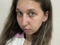 Sanella has a serious face with light green eyes, slightly wavy brown hair, and a small nose. She's wearing a pink and white shirt with a pearl necklace.