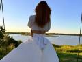 MiraGi, con lunghi capelli rossi, un top bianco a vita corta e una gonna bianca con un grande fiocco, è in piedi su un'altalena, rivolta verso l'acqua, con il sole al tramonto e un cielo sereno. Le sue natiche sono parzialmente visibili..