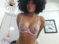 SnackTime with curly black hair, light brown skin, wearing pink lace bra with floral embroidery, showing her small tits, standing in a room with a gold pendant necklace.