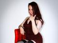 SweettEve with red hair, smiling, wearing a dark red dress, black thigh-highs, and red nails, sitting on a red chair, giving a thumbs-up, with her finger on her lip.
