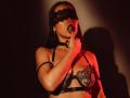 JaneWow with black lace blindfold, biting finger, wearing black lace bra with straps, choker, and arm band, red light on her, shadow on wall, looking seductive.