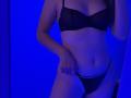 Alice, vêtue de lingerie noire, expose ses seins et ses parties intimes dans un soutien-gorge et une culotte transparents, avec un fond éclairé en bleu, une main sur sa hanche, et un piercing au nombril visible..
