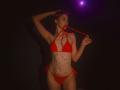 AgataRyili with small red bikini, holding string in mouth, body covered in colorful star tattoos, dark background, purple spotlight, slim, fair-skinned, blonde, hand in hair.