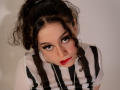 KendraSimons with curly black hair in a ponytail, wearing a striped black-and-white shirt, looking up with red lipstick, long eyelashes, and a nose ring, in a photo.