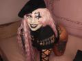 Chili with pink hair, black beret, white makeup, black lipstick, smiling, wearing black fishnet top, exposing tattoos, on pink bed, knees on pillow, showing ass, tattooed thighs.