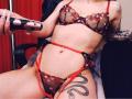 HotCaramelsTrio with pale skin, wearing sheer black and red floral lingerie, holding a black dildo, tattoo of a snake on right thigh, red garter straps, sitting on chair.
