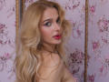FloraDavis has long, wavy blonde hair and is looking over her shoulder with green eyes. She has pink lipstick and light makeup. Her right shoulder is slightly exposed. The background is pink floral wallpaper with gold trim. The photo focuses on her face and upper body. Her hair is soft and slightly messy. The floral pattern is detailed and adds a vintage feel. FloraDavis's expression is slightly curious. The image is sharp and well-lit.