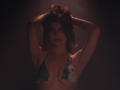 StellaLewin with her arms raised, wearing a blue and white tie-dye bikini top, dark brown wavy hair, and a subtle necklace, in a dimly lit, moody setting.