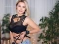 BelleLisaG with blonde bob, wearing black mesh top, showing tits, black strappy harness, denim shorts, and black wristbands, standing in front of green plants.