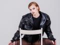 GrettaDemichelis with red hair, black leather jacket, black top, black tights, sitting on a white chair, looking at camera with a slight smirk, hands on chair's seat.