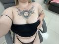DiLaria with sun tattoo on chest, black strapless top, black thong, sitting in chair, showing off her big tits and belly, blonde hair, yellow nails, white tile floor.
