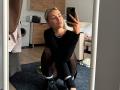 ArnettaNazario squatting, taking a mirror selfie, wearing a black long-sleeve top, black tights, black boots, and a pearl necklace, with pouty lips, blonde hair, and a ring on her finger.