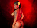 TamaraLubezki, nude, stands with her back to the camera, head turned to smile at the viewer. She has dark skin, a tattoo on her right arm, and a bright orange flower in her hair. Her breasts are visible from the side, and she has a curvy, well-defined buttocks. The red, textured background contrasts with her skin tone. Her smile is confident and playful. The photograph emphasizes her natural beauty and body shape. The lighting highlights her skin and the tattoo.