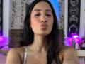 JennTylor with pouty lips, dark hair, and subtle makeup, wearing a grey top, showing a small tattoo on her right shoulder. Her pussy is not visible.