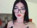 IsabellaaRossi with black hair, glasses, and pink lipstick, showing off her tits in a grey strap top. Her ass is not visible. She's looking directly at the camera.