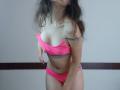 VikkiRi with wavy brown hair, wearing a pink bikini that's slightly slipping to reveal her small tits and gray underwear, leaning forward with a slight smile.