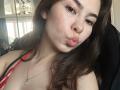 EmeraldCrivaro with puffy pink lips, brown wavy hair, and a red and black strap top, wearing a silver necklace, has a cute, slightly chubby face, and brown eyes.