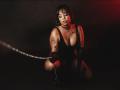 MadisonHetfield kneels in black lingerie, holding a chain with gloved hands. She has tattoos on her arm, dark makeup, and a choker. Her tits are visible, with red lighting.