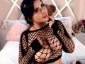 AgathaVega with black fishnet top, exposing her large tits, dark hair, and pouty expression, touching her chin in a bedroom with a pink pillow and teddy bear.