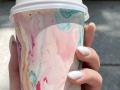 Eine Hand hält ein Eisbecher mit rosa-blauen Wirbeln, einem weißen Deckel, rosa Nägeln und einer weißen Plastikhülle, auf einer grauen, strukturierten Oberfläche..