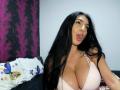 BrunetteBabe69 with long black hair, pouty lips, and big tits in a pink checkered bra, looking at the camera. Purple butterfly pillow in the background.