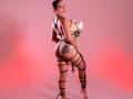 JessicaFiore, con un sedere importante, che tiene in mano rose bianche, indossa un intimo nero con spalline sottili, ha un tatuaggio sulla coscia destra, illuminazione rossa e rosa, capelli scuri raccolti in uno chignon, guarda oltre la spalla..