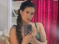 HannaRobertss with long brown braid, green lace top, holding phone with purple case, red curtains, and white shelves in background, looking at camera with subtle smile.