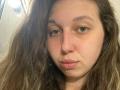 Sanella has wavy brown hair, green eyes, and a slightly pouty face with pink lips. She's looking at the camera with a neutral expression. Her nose is small.