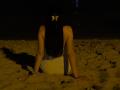 Olieri is on a sandy beach at night, wearing a white dress, with long black hair and a butterfly hairclip, sitting with arms on sand, back to camera.