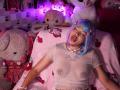 LiMiuu with blue and pink bob, eyes closed, tongue out, wearing sheer pink top, exposing nipples, white necklace, and pink bracelet, surrounded by stuffed animals, in pink-lit room.