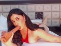 VickyMegan with long black hair, red bikini, lying on bed, sucking a large, uncircumcised dildo, her tits slightly visible, her ass raised, legs crossed behind.