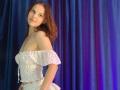 ChayaParrett with red curly hair, white off-shoulder floral top, and matching skirt, standing sideways, smiling, showing small tits, against blue and purple draped fabric.