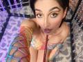 LunaixMoon with colorful fishnet arm sleeves, looking up with purple lipstick, holding a red leash, wearing a colorful off-shoulder top, in a room with purple and white patterns.