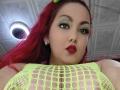 MirandaRossi with bright red hair, green eye makeup, red lipstick, wearing a neon yellow fishnet top, showing her large, round tits. Ceiling light in background.