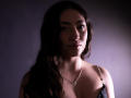 AnnFisher has long dark brown hair, a slim face, and is wearing a necklace with a small pendant. She's in a dark, softly lit, upper-body shot. Boobs visible.