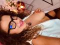 BustyMira with curly brown hair, blue eyes, and glasses, smiling, topless, showing tits, red nail polish, white shirt, black panties, and a necklace. Bedroom setting.