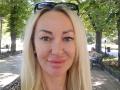 MysteryLaddy has blonde hair, green eyes, and a small nose with pink lips, wearing black sunglasses on her head. She's smiling in a park with trees and a bench.