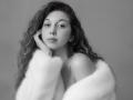Sanella with wavy hair, wearing a fluffy white coat, hand on cheek, looking slightly to the side, gray background, black and white, soft expression, bare shoulders, subtle beauty.