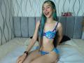 AbbyHillsI, with green and blue hair, sits on a bed wearing a blue floral bra and panty set, smiling with her mouth open, showing her teeth. Her tits are small.