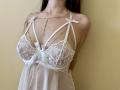 Tisia with big tits wearing a white lace babydoll, ribbons, and silver chain necklace, her arms behind her back, long dark hair, and a pale yellow wall.