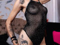 LunaLindaHot wearing a black mesh, sparkly one-piece that reveals her small, firm tits and tattoos on her arm and thigh, standing in a bedroom.